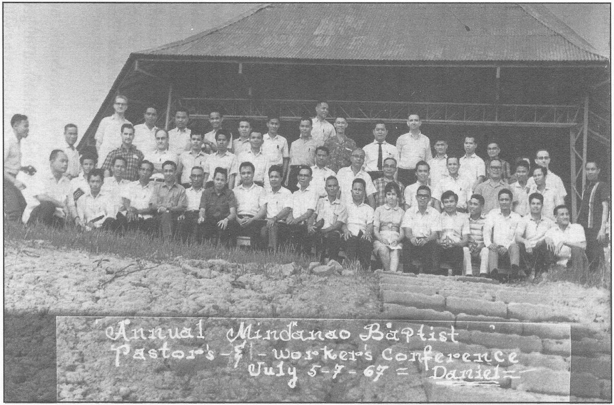 Large gathering of pastors and church workers posed outside a building with a hand-lettered banner.
